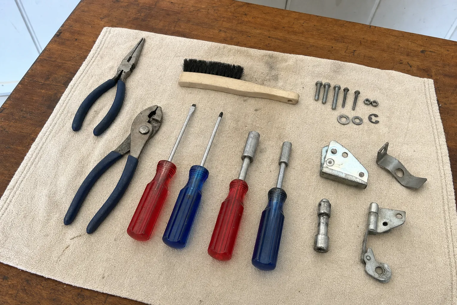 Two pairs of pliers, red and blue screwdrivers and nut drivers on a canvas cloth beside a pivot bracket with a pin barrel, a mounting plate, a sleeve and bolts